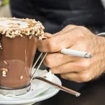 Uomo seduto al PC con un dettaglio della mano con una sigaretta e un bicchiere di cioccolata