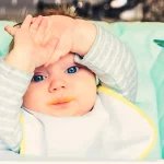 Bambino seduto su un seggiolone con le mani sulla fronte che sembra voler rifiutare la pappa