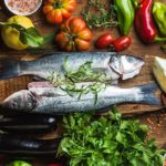 Tavolo con alimenti sani: 2 spigole, peperoni, limone, melanzane, prezzemolo, spinaci aglio, olio, pomodori e spezie