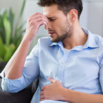 Ragazzo seduto su un divano con una mano sulla pancia e una sul naso con espressione del viso dolorante